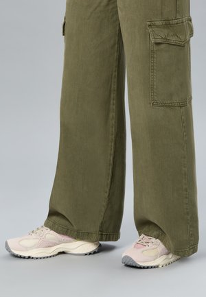 Pantalon cargo vert olive à jambes larges porté avec des baskets épaisses rose clair et crème sur un fond gris uni.