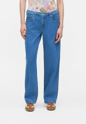 Personne portant un jean bleu droit, un haut fleuri rentré à l’intérieur et des baskets marron avec des lacets rouges, debout devant un fond uni.