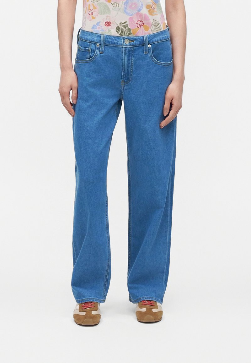 Personne portant un jean bleu droit, un haut fleuri rentré à l’intérieur et des baskets marron avec des lacets rouges, debout devant un fond uni.