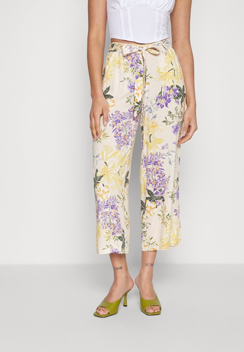 Femme portant un pantalon floral court avec des fleurs jaunes et violettes, un haut court blanc et des sandales à talons olive, se tenant sur un fond uni.
