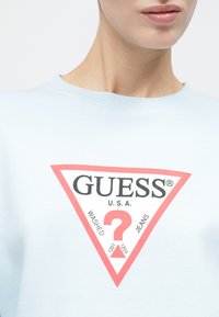 Κοντινό πλάνο ενός ατόμου που φοράει ένα ανοιχτό μπλε πουκάμισο με κόκκινο και λευκό λογότυπο Guess που περιλαμβάνει ερωτηματικό και κείμενο.