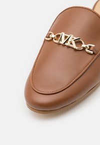 Mocassin en cuir marron avec détail en chaîne dorée et logo "MK" sur le dessus, présentant une texture lisse et un design à bout arrondi.