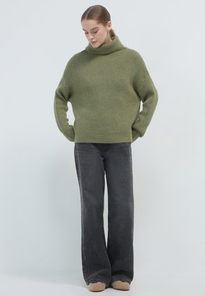 Pull en tricot vert avec un col montant, associé à un jean large gris foncé. La texture est douce et épaisse, offrant une coupe décontractée.