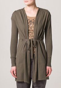 Olivgrön kofta med draperad front och knytband i midjan. Långa ärmar, mjukt tyg och avslappnad passform. Bärs över en mönstrad beige topp.