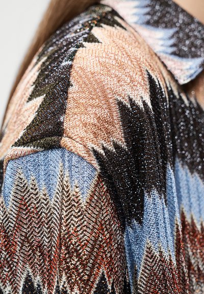 Κοντινό πλεκτού υφάσματος με ζιγκ-ζαγκ μοτίβα σε μπεζ, μαύρο, μπλε, κόκκινο και λευκό στον ώμο ενός ατόμου.