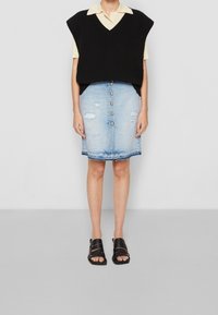 Zwarte gebreide vest over een lichtgele blouse met korte mouwen en kraag. Lichtblauwe distressed spijkerrok met knoopsluiting aan de voorkant en zakken. Zwarte sandalen.
