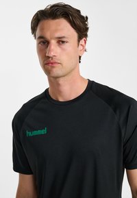 Czarny sportowy t-shirt z krótkimi rękawami, okrągłym dekoltem i zielonym logo „hummel” na lewej piersi. Gładka, lekka tkanina.