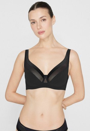 Žena s tmavými vlasy v černé podprsence a kalhotkách stojí před jednoduchým světlým pozadím s rukama za zády.