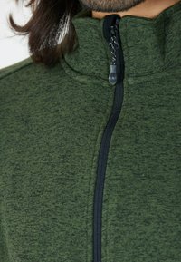 Giacca in poliestere verde con zip, caratterizzata da un colletto alto, una zip nera, un tessuto testurizzato e un sottile motivo melangiato.