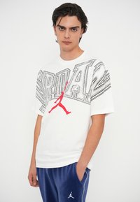 Fehér pamut póló nagy fekete és piros grafikával, sziluettel, kék bársony melegítőnadrággal, logó részlettel párosítva.