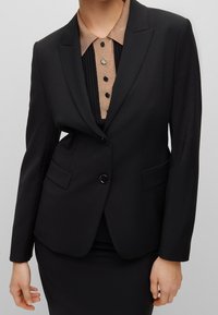 Mustat bleiserit, joissa on huippulaput, yksittäinen nappisulku, kaksi sivutaskua, yhdistettynä kaulukselliseen laskostettuun puseroon, jossa on mustat napit.