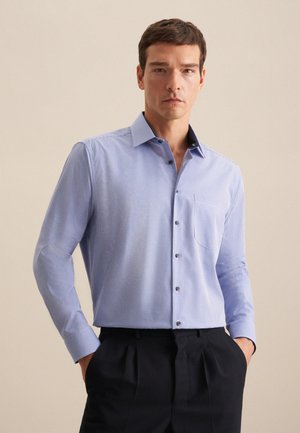 Homme portant une chemise à manches longues à carreaux bleu clair rentrée dans un pantalon plissé noir, debout les mains dans les poches devant un fond beige.