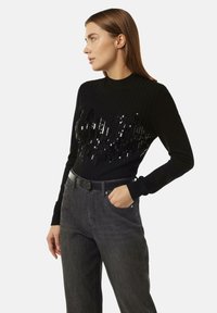 Schwarzer gerippter Pullover mit glitzerndem Design auf der Vorderseite, langen Ärmeln, hohem Kragen, kombiniert mit dunklen Jeans und einem eleganten schwarzen Gürtel.