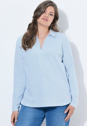 Femme aux longs cheveux bruns portant une chemise à manches longues rayée blanche et bleu clair et un pantalon bleu, debout devant un fond blanc uni.