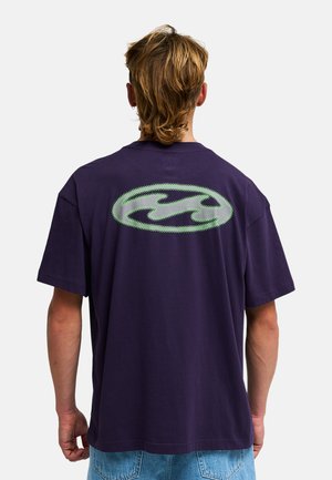 Person mit mittellangen braunen Haaren trägt ein dunkelviolettes T-Shirt mit einem grünen ovalen Motiv auf dem oberen Rücken und hellblaue Jeans.