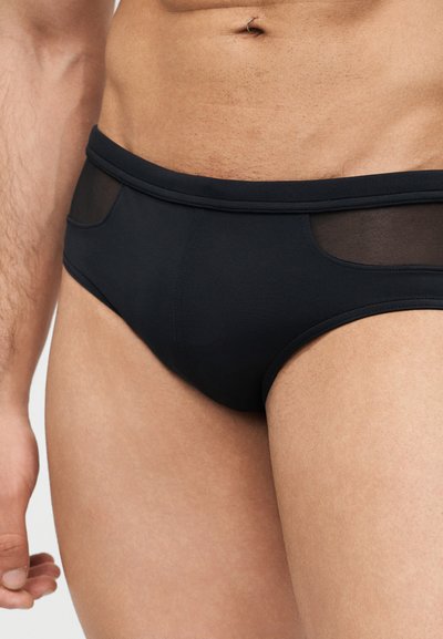 Κοντινό πλάνο ενός ατόμου που φοράει μαύρο εσώρουχο τύπου σλιπ με ημιδιαφανή πλευρικά πάνελ από δίχτυ και εμφανές τρύπημα στον αφαλό.