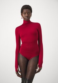 Červený bodysuit s dlhým rukávom a vysokým golierom, vyrobený z hladkého materiálu; nosený s priesvitnými pančuchami s bodkami a čižmami po kolená.