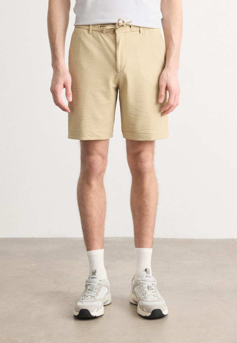 Shorts de color beige con textura, cintura elástica con cordón, bolsillos frontales y diseño ajustado, combinados con calcetines blancos y zapatillas grises.