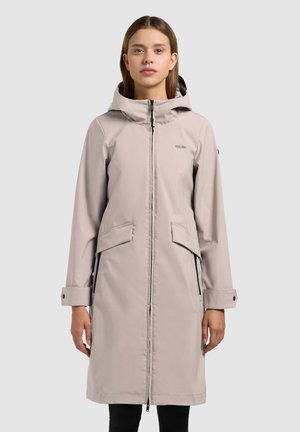 Kvinde iført en lys beige knælængde hættejakke med lynlås foran og klaplommer, stående foran en ensfarvet baggrund.