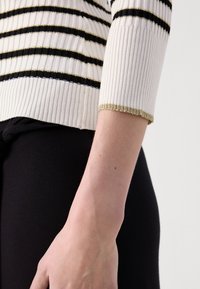 Pull en maille blanche à rayures noires et dorées, avec des manches trois-quarts ajustées et un poignet rehaussé d'or. Tissu noir en dessous.
