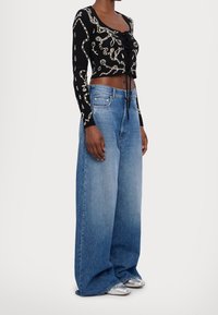 Femme portant un haut noir crop top à manches longues avec des motifs beige, un jean à jambes larges bleu et des chaussures argentées à bouts pointus, debout devant un fond blanc.
