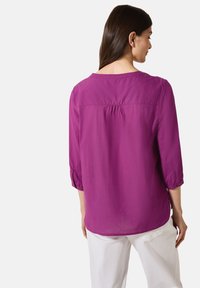 Lila, leichtes Blusenshirt mit gerafftem Detail auf der Rückseite, rundem Ausschnitt und hochgekrempelten Ärmeln. Kombiniert mit weißen Hosen.