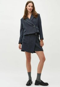 Donkergrijze, op maat gemaakte blazer met dubbele knopen, gecombineerd met een bijpassende rok. Gedragen met glanzende zwarte schoenen en grijze sokken. Gladde textuur.