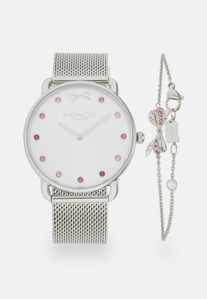Reloj con correa de malla plateada y esfera blanca, que presenta piedras rosas como marcadores de hora y un acento en forma de lazo. Incluye una pulsera a juego con charms.