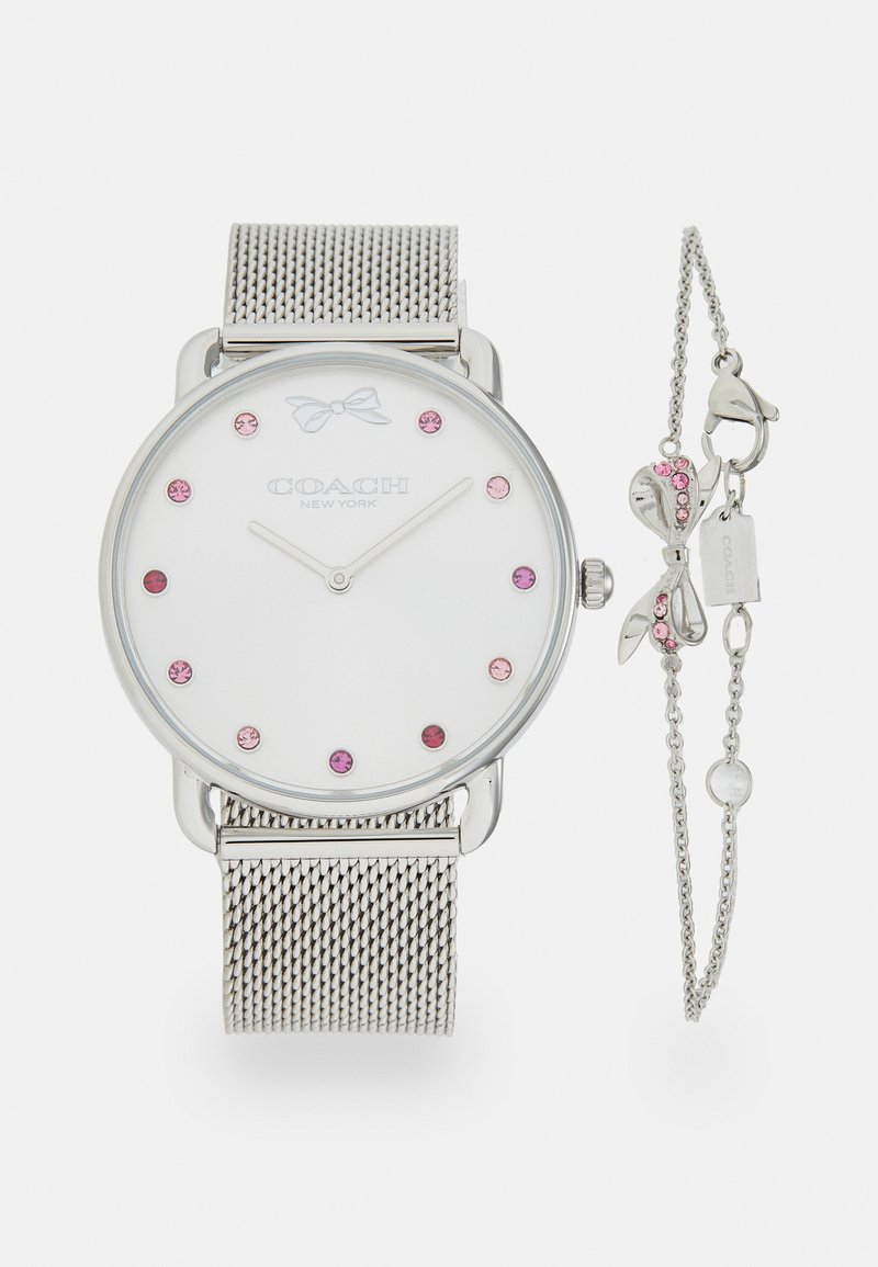 Hõbedane võrksangaga kell valge näo ja roosa kividest tunni indikaatoritega ning väikese paela aktsendiga. Kaasas on sobiv ehtekäevõru, milles on ripatsid.