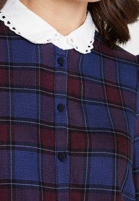 Plaid shirt featuring dark blue, burgundy, and light blue tones; white collar with scalloped edge; front buttons for closure.
