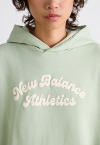 Felpa con cappuccio verde chiaro caratterizzata da lettere bianche in rilievo che recitano "New Balance Athletics". Tessuto morbido con vestibilità rilassata e una leggera trama.