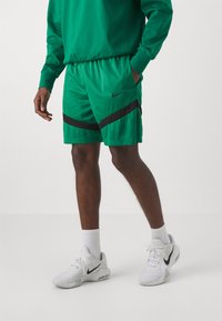 Žalios tinklinės šortai su juodais įstrižais raštais, elastinga juostos dalimi ir šoninėmis kišenėmis. Dėvimi su baltais Nike sportbačiais ir baltomis kojinėmis.