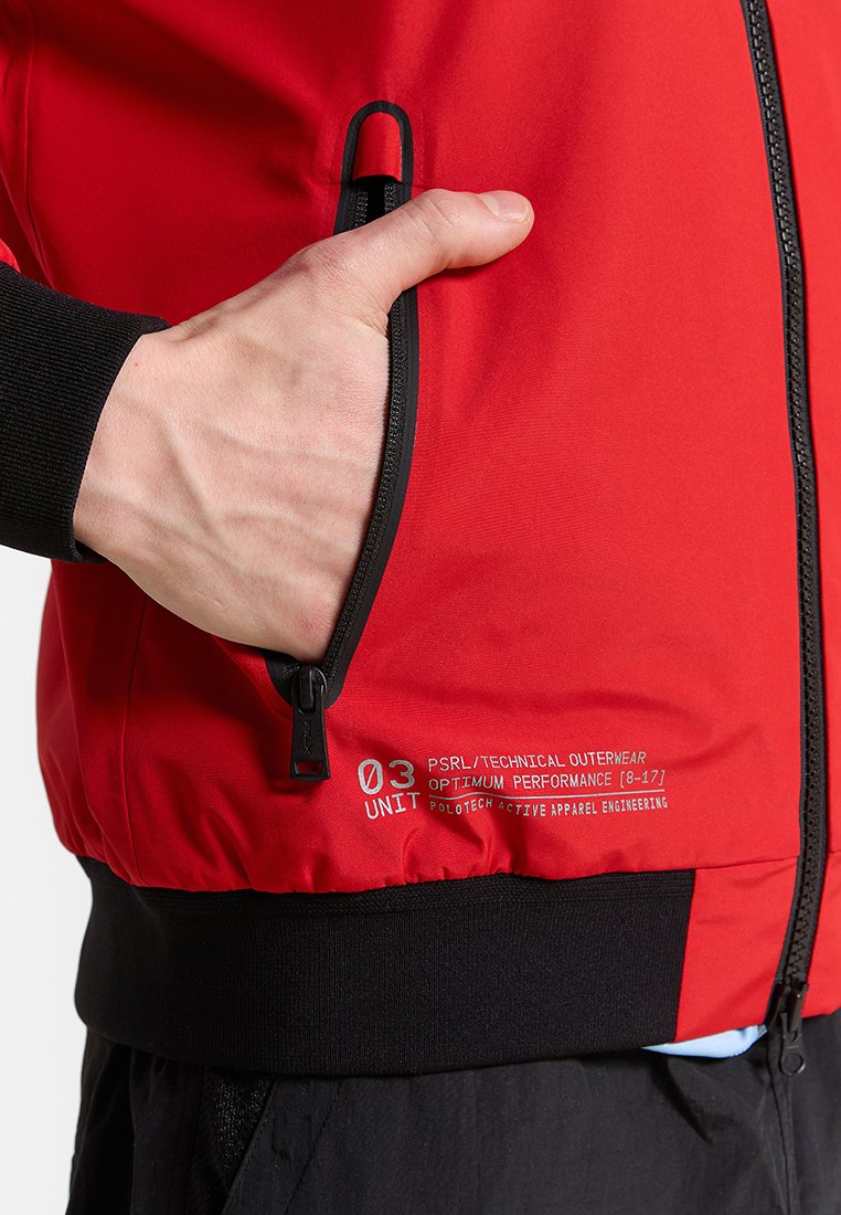 Chaqueta exterior técnica roja con un bolsillo con cremallera, acentos negros y una marca etiquetada. Textura suave con un diseño aerodinámico.
