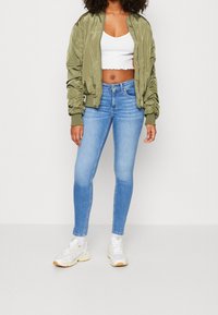 Olivově zelená bomber bunda, bílý kostkovaný top, modré skinny džíny a bílé tenisky. Hladká tkanina s vroubkovanými manžetami a pasem.