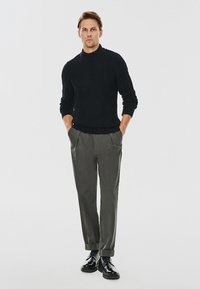 Czarny sweter w splot warkoczowy, szare spodnie i czarne buty. Sweter ma wysoki kołnierz; spodnie mają luźny krój z kantem.
