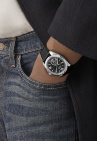 Reloj plateado con correa de cuero negro, caja redonda y esfera con números romanos, que presenta un movimiento esqueleto para visibilidad de los mecanismos interiores.