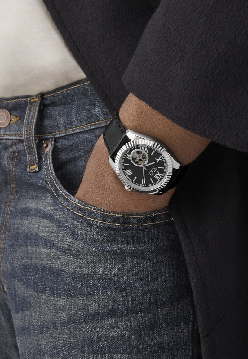 Reloj plateado con correa de cuero negro, caja redonda y esfera con números romanos, que presenta un movimiento esqueleto para visibilidad de los mecanismos interiores.