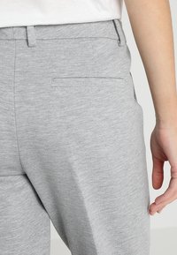 Pantaloni grigi con una texture liscia, dotati di una tasca posteriore e di una vita, mostrati di lato con parte di una mano visibile.
