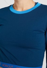 Long-sleeve top in dark blue with a light blue neckline, featuring a textured fabric and a waistband that reads "WORKOUT" in orange.