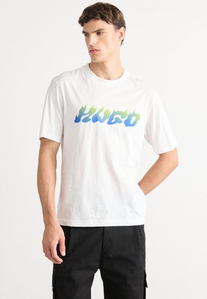Young man wearing white T-shirt with blue and green flame-style "Hugo" logo, standing with hand in pocket, looking to side.