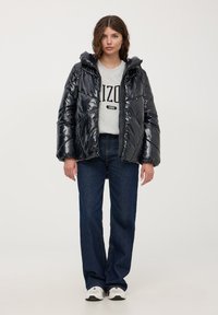 Giacca in nylon nera imbottita con cappuccio, caratterizzata da una texture a rombi. Indossata sopra un felpa grigia e abbinata a jeans blu scuro.