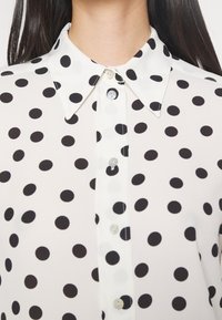 Chemise blanche boutonnée avec des pois noirs portée par une personne aux longs cheveux foncés, montrant le col et le haut du torse.