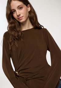 Top lungo marrone a maniche lunghe con scollatura rotonda, caratterizzato da un design attorcigliato e un accento circolare in vita. Tessuto morbido con una superficie liscia.