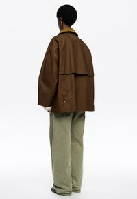 Hnedý oversized kabát s ventilačným chrbtom, kontrastným horčicovým golierom a voľnými zelenými nohavicami, dokončujúci uvoľnený outfit.