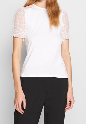 Mujer con una blusa blanca con mangas abullonadas transparentes con lunares y pantalones negros sobre un fondo liso y claro.