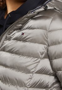 Lichtgrijze pufferjack met een gestikte design, glanzende textuur en een klein geborduurd logo. Heeft een capuchon en een ritssluiting.