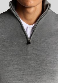 Pullover grigio a metà zip realizzato in tessuto morbido, con una texture liscia e un tirante della zip in contrasto, abbinato a una camicia bianca con colletto sotto.