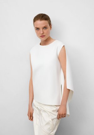 Haut sans manches blanc avec un col rond, un design asymétrique et une texture lisse. Associé à un pantalon slim drapé de couleur claire.