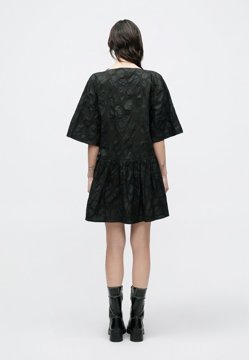 Robe noire à manches courtes, taille resserrée et tissu texturé. Portée avec des bottines noires à fermeture éclair latérale.