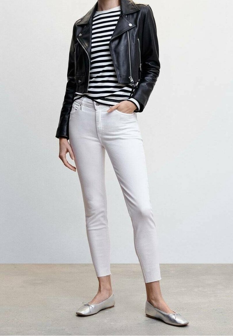 Personne portant une veste en cuir noire, une chemise rayée noire et blanche, un pantalon blanc ajusté et des ballerines argentées, debout avec une main dans la poche.